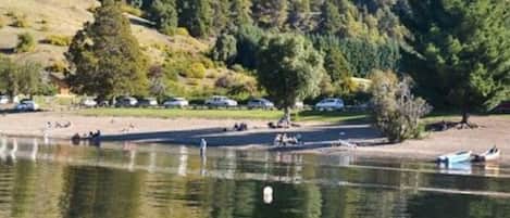 Un lago tranquilo con una colina boscosa al fondo, algunas embarcaciones y gente cerca de la orilla.