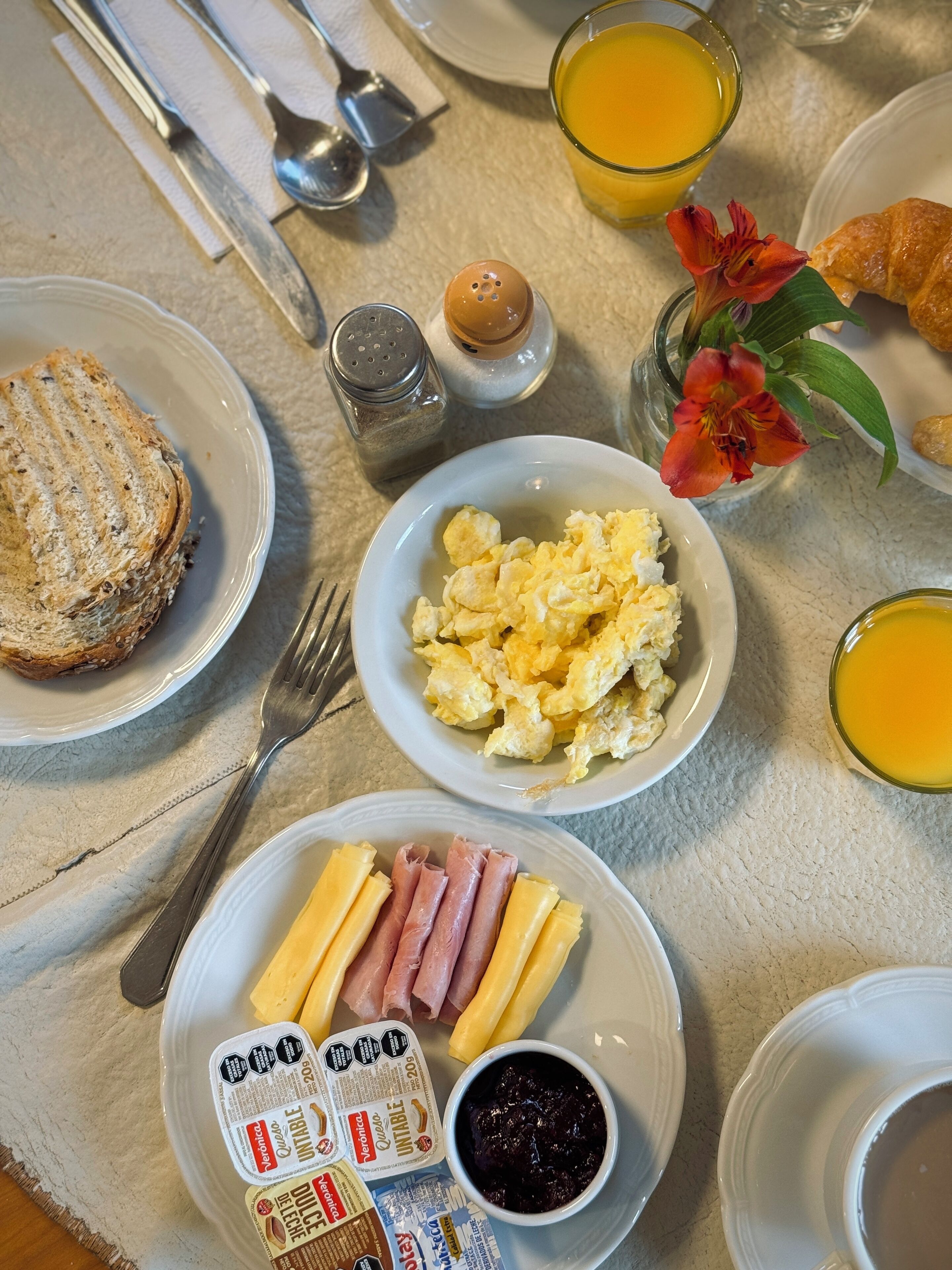 A breakfast table with plates of scrambled eggs, ham, cheese, croissant, and toast. There are also two glasses of orange juice and a flower in a vase.