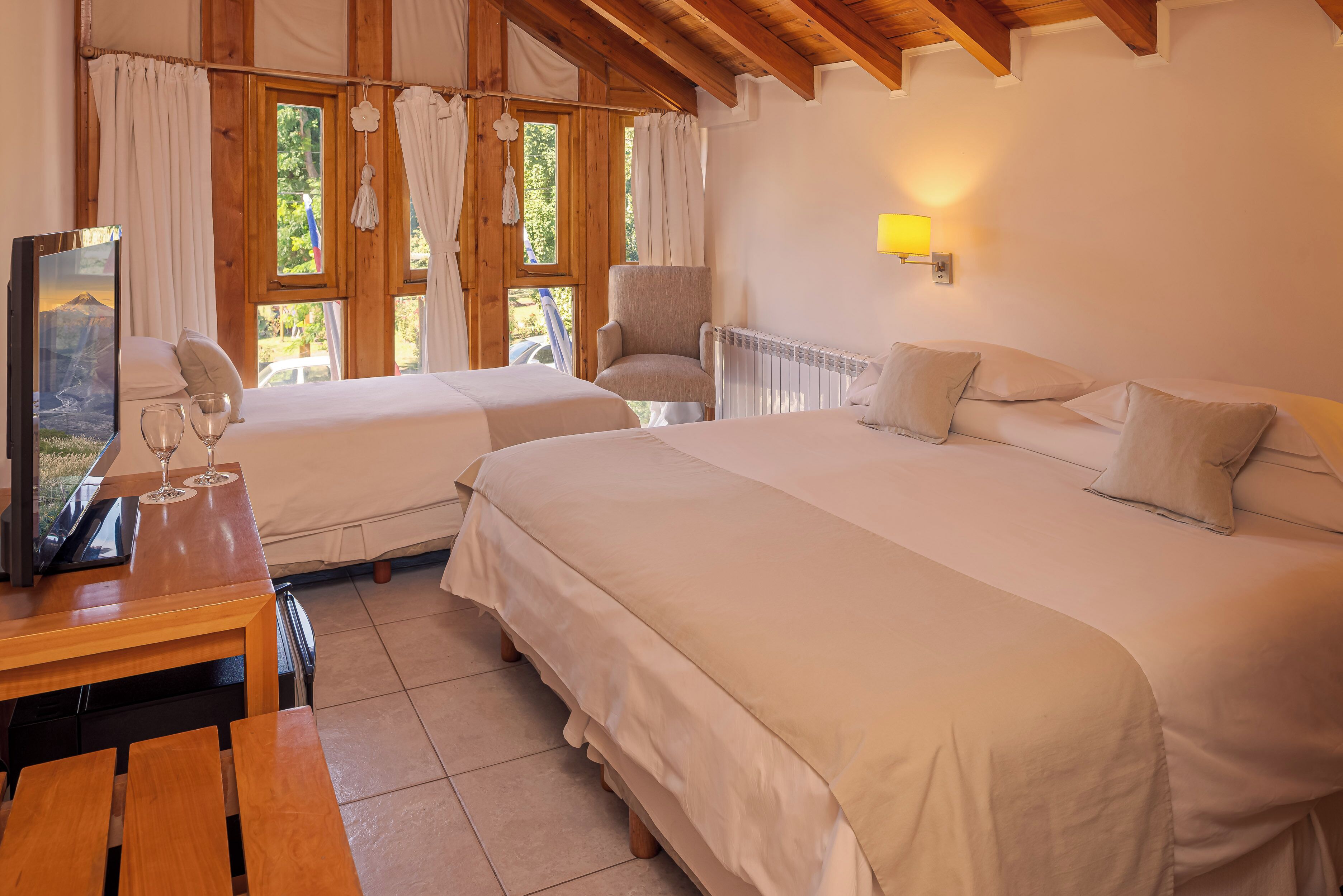 A room with two beds, a wooden table with a TV, and a window with curtains.