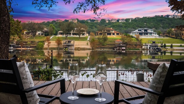 Un patio junto al lago con dos copas de vino vacías sobre una mesa, rodeado de sillas, con vista a un lago y casas y árboles al fondo.