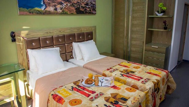 A hotel room with a bed, a glass table, a wooden wardrobe, and a painting of a coastal town. - Wakola business hotels (Dakar)