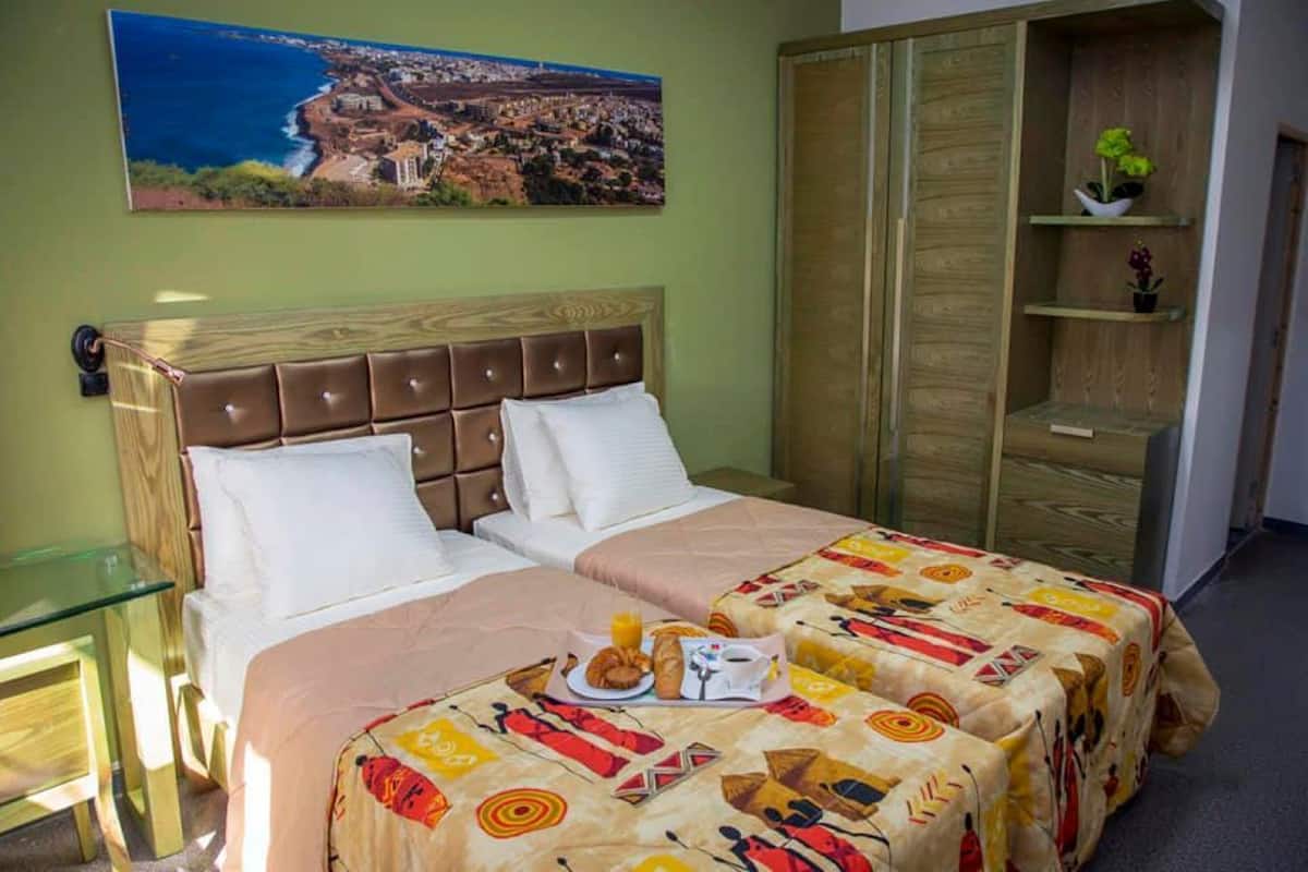 Une chambre d’hôtel avec un lit, une table en verre, une armoire en bois et un tableau représentant une ville côtière.
