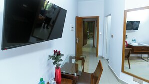 A hotel room with a flat-screen TV mounted on the wall, a wooden dining table with chairs, a glass coffee table, and a red vase with flowers. - Wakola business hotels (Dakar)