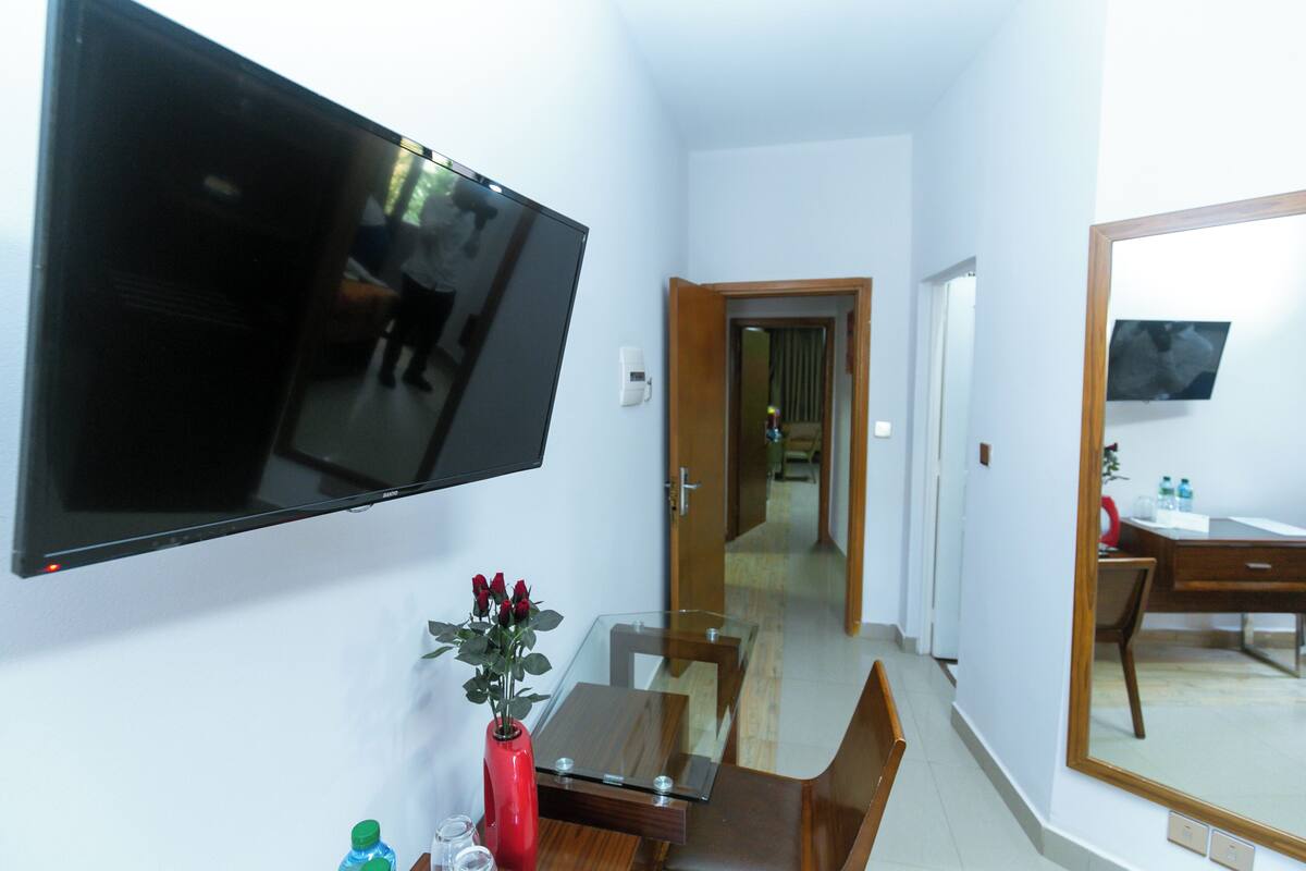 Une chambre d’hôtel dotée d’un téléviseur à écran plat fixé au mur, d’une table à manger en bois avec des chaises, d’une table basse en verre et d’un vase rouge contenant des fleurs.