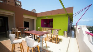 An outdoor patio with a dining area featuring wooden tables and chairs, a small bar area with a purple counter, and a view of the surrounding area. - Wakola business hotels (Dakar)