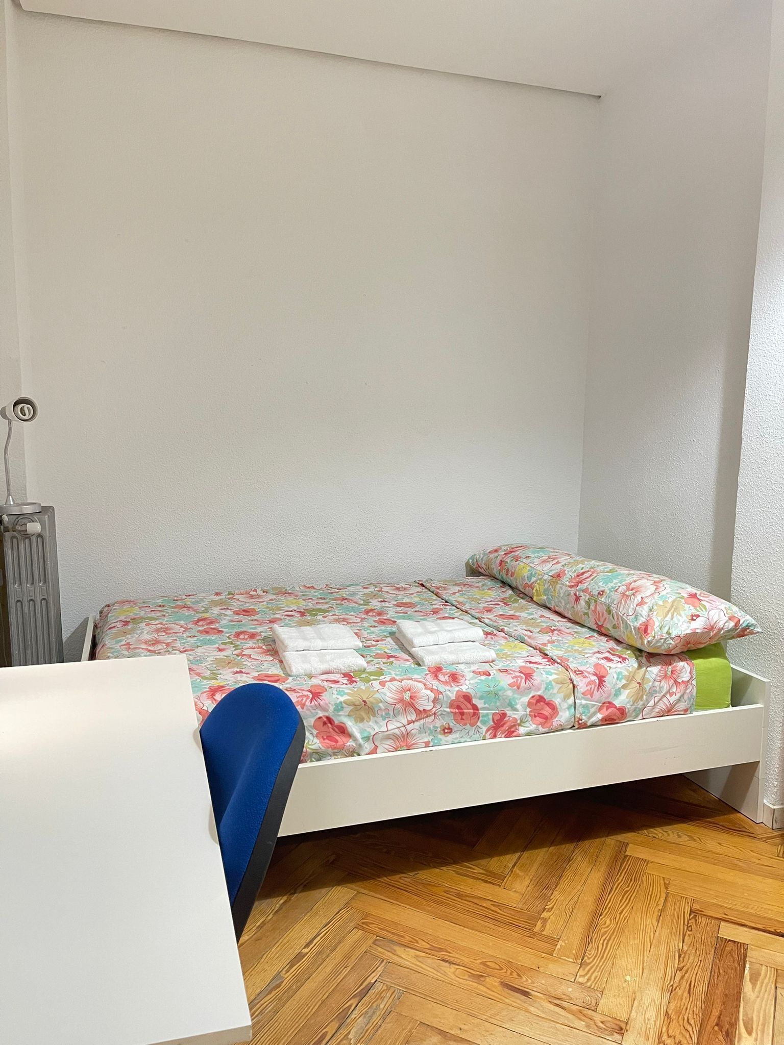 Une petite chambre avec un lit simple, un plancher en bois, une tête de lit blanche et une couvre-lit à motifs floraux.