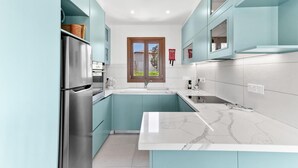 Una cocina moderna con armarios color turquesa, un refrigerador de acero inoxidable, una encimera de mármol blanco y una ventana con vistas a la vegetación.