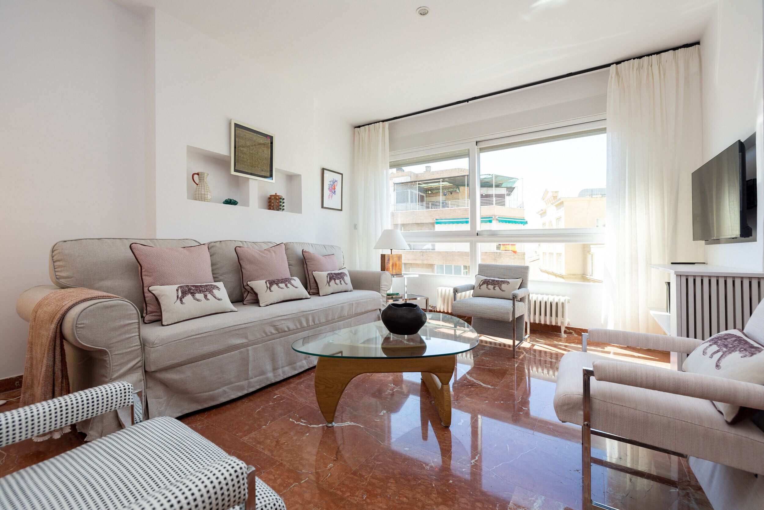 A modern living room with a glass coffee table, a beige sofa with patterned cushions, a striped armchair, and a view of a balcony with a pool.