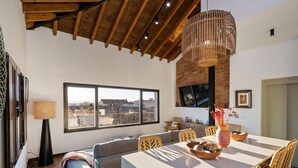 A modern living room with a large window, a dining area with wicker chairs, and a wooden ceiling. - ChezmoiHomes Santiago Duplex Penthouse (Granada)