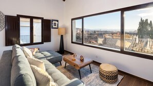 A living room with a sofa, a coffee table, a lamp, and a view of the city through large windows. - ChezmoiHomes Santiago Duplex Penthouse (Granada)