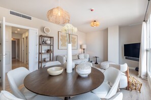 A modern living room with a round dining table, white chairs, a large vase with flowers, and a flat-screen TV mounted on the wall.