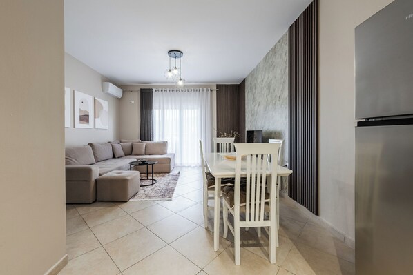 A modern living room with a sectional sofa, a dining table with chairs, a pendant light, and a large window with sheer curtains.