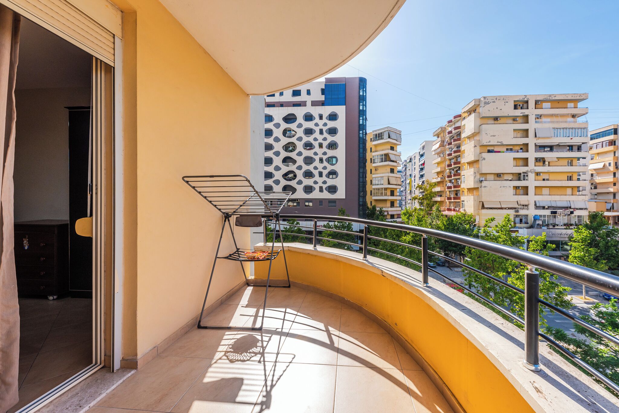 A balcony with a metal chair and a drying rack, overlooking a cityscape with modern buildings.