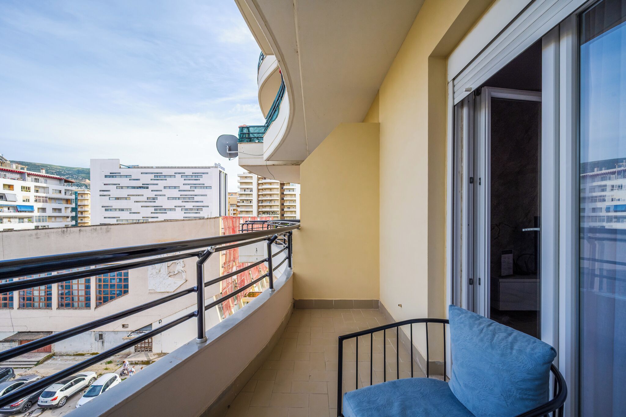 A balcony with a blue chair, a view of buildings, and a satellite dish.