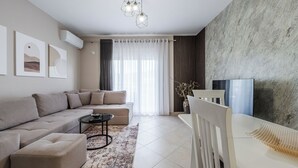 A modern living room with a sectional sofa, a coffee table, a white armchair, and a TV on a stand. - Kondo Stays - City Center Apartments (Vlorë)