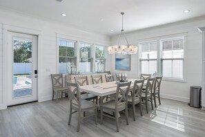 A dining area with a large table, multiple chairs, a chandelier, and large windows with blinds. - Seaspray: Pool! Sleeps 26! Beach access! (Destin)