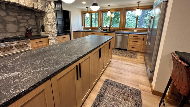 A modern kitchen with a granite countertop, wooden cabinets, a stone accent wall, and large windows.