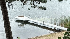 A wooden dock extending into a calm lake, surrounded by trees and a stone pathway leading to a sandy beach.