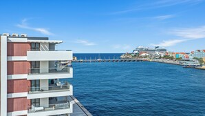 Ein Hotel am Wasser mit mehreren Balkonen, einer Pier mit Booten und einem Kreuzfahrtschiff im Hintergrund.