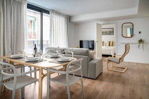 A modern living room with a dining area, a grey sofa, a TV, and a wooden floor.