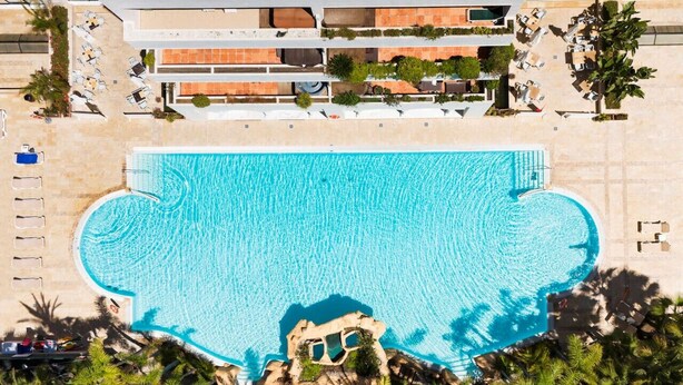 An aerial view of a hotel with a large swimming pool, multiple lounge areas, and a well-maintained garden.