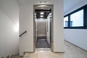 A modern elevator with a metallic finish, a handrail on the left, and a clear glass door.