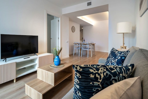 A modern living room with a flat-screen TV, a sofa with patterned cushions, a coffee table, and a dining area with a table and chairs.