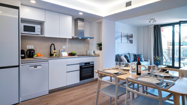 A modern kitchen with white cabinetry, a built-in microwave, and a dishwasher. There is a dining area with a wooden table and chairs, and a window with a view of the city. - Habitat Apartments Teatre 22 (Barcelona)
