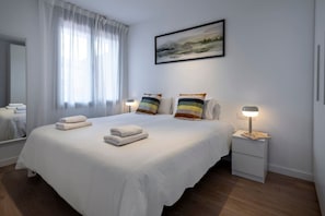 A hotel room with a large bed, two bedside tables, a nightstand, a lamp, a painting, and a window with sheer curtains.