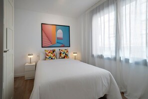 Ein Hotelzimmer mit einem Bett, zwei Nachttischlampen, einem farbenfrohen abstrakten Gemälde und durchscheinenden Vorhängen.