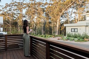 Une personne se tient sur une terrasse en bois, d’où l’on aperçoit un jardin avec des arbres et un bâtiment moderne.