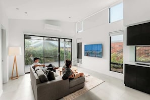 Ein modernes Wohnzimmer mit einer Couch, einem Couchtisch und einem Fernseher. Große Fenster und Blick ins Grüne sind zu sehen.