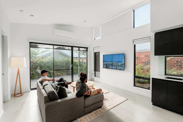 Ein modernes Wohnzimmer mit einer Couch, einem Couchtisch und einem Fernseher. Große Fenster und Blick ins Grüne sind zu sehen.