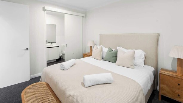 Une chambre d’hôtel avec un lit, une table de chevet en bois, une chaise, une salle de bain avec un miroir et une porte.