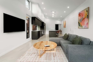 Ein modernes Wohnzimmer mit Flachbildfernseher, grauem Sofa, Holztisch und einer Küche im Hintergrund.