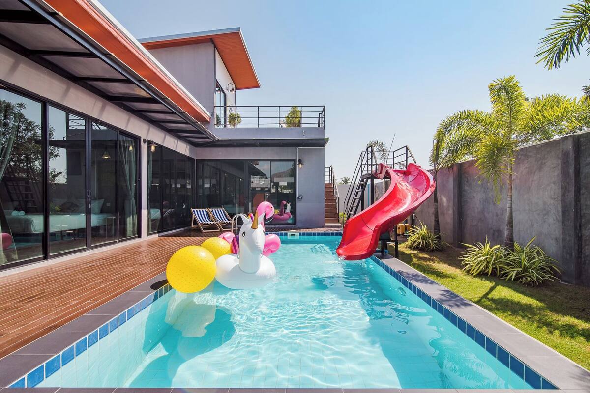 A modern house with a swimming pool, a red slide, and inflatable unicorn and flamingo toys.