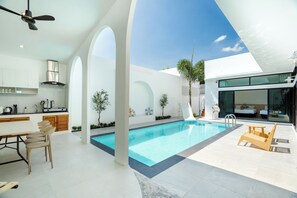 Et moderne køkken med hvide skabe, et spiseområde med træstole, en swimmingpool og et opholdsområde med træmøbler.