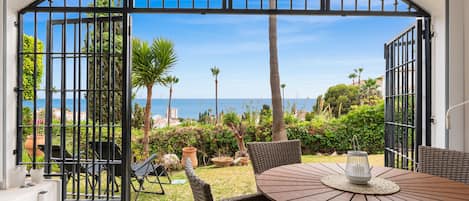 An outdoor patio with a round table and wicker chairs, overlooking a garden with palm trees and a view of the sea.