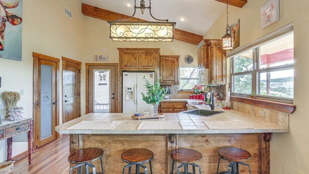 A spacious kitchen with wooden cabinets, a large island with stools, and a view through the window. - Medicine Park Cottage: Walk to Lake Lawtonka! (Medicine Park)