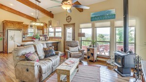 A cozy living room with a fireplace, a sofa, a dining area, and a kitchen with wooden beams and a ceiling fan. - Medicine Park Cottage: Walk to Lake Lawtonka! (Medicine Park)
