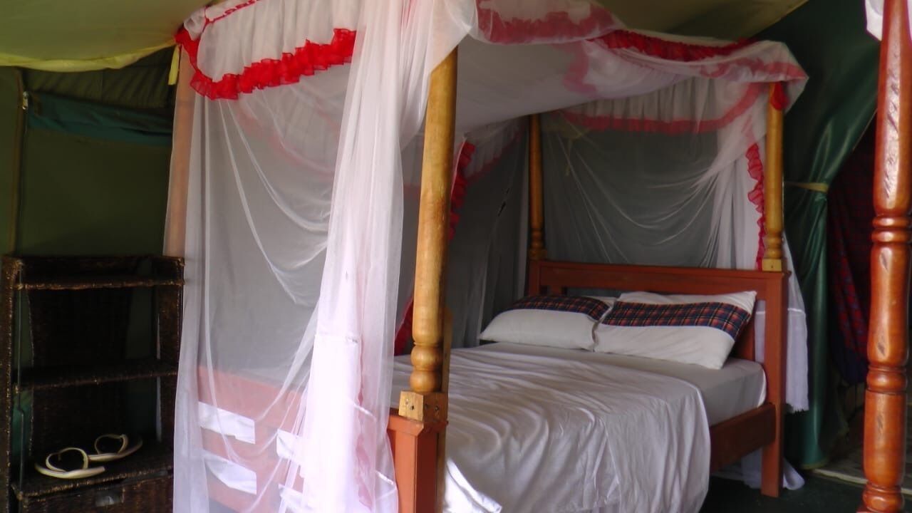 Una cama con dosel y cuatro columnas, ropa de cama blanca con detalles rojos, un cabecero de madera y una mesita de noche con lámpara.