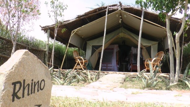 Une structure rustique en forme de tente, dotée d’un toit de chaume, entourée de chaises et d’une pierre sur laquelle est gravé le mot « Rhino ».
