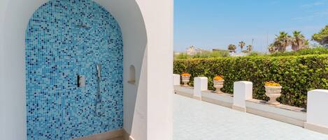 Un área de piscina con baldosas, una pared de mosaicos azules, cielo despejado y un seto al fondo.