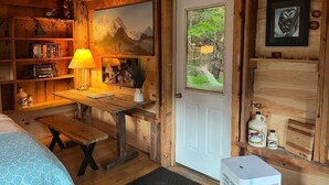 A cozy cabin room with wooden walls, a bed, a desk with a lamp, a bookshelf, and a door leading outside. - Expedition Everest Cabin At Basecamp (Saint Johnsbury)