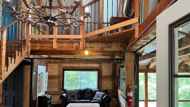 A spacious log cabin interior with a high ceiling, a large chandelier, a cozy seating area with a sofa and ottoman, and a view of the outdoors through large windows. - Expedition Everest Cabin At Basecamp (Saint Johnsbury)