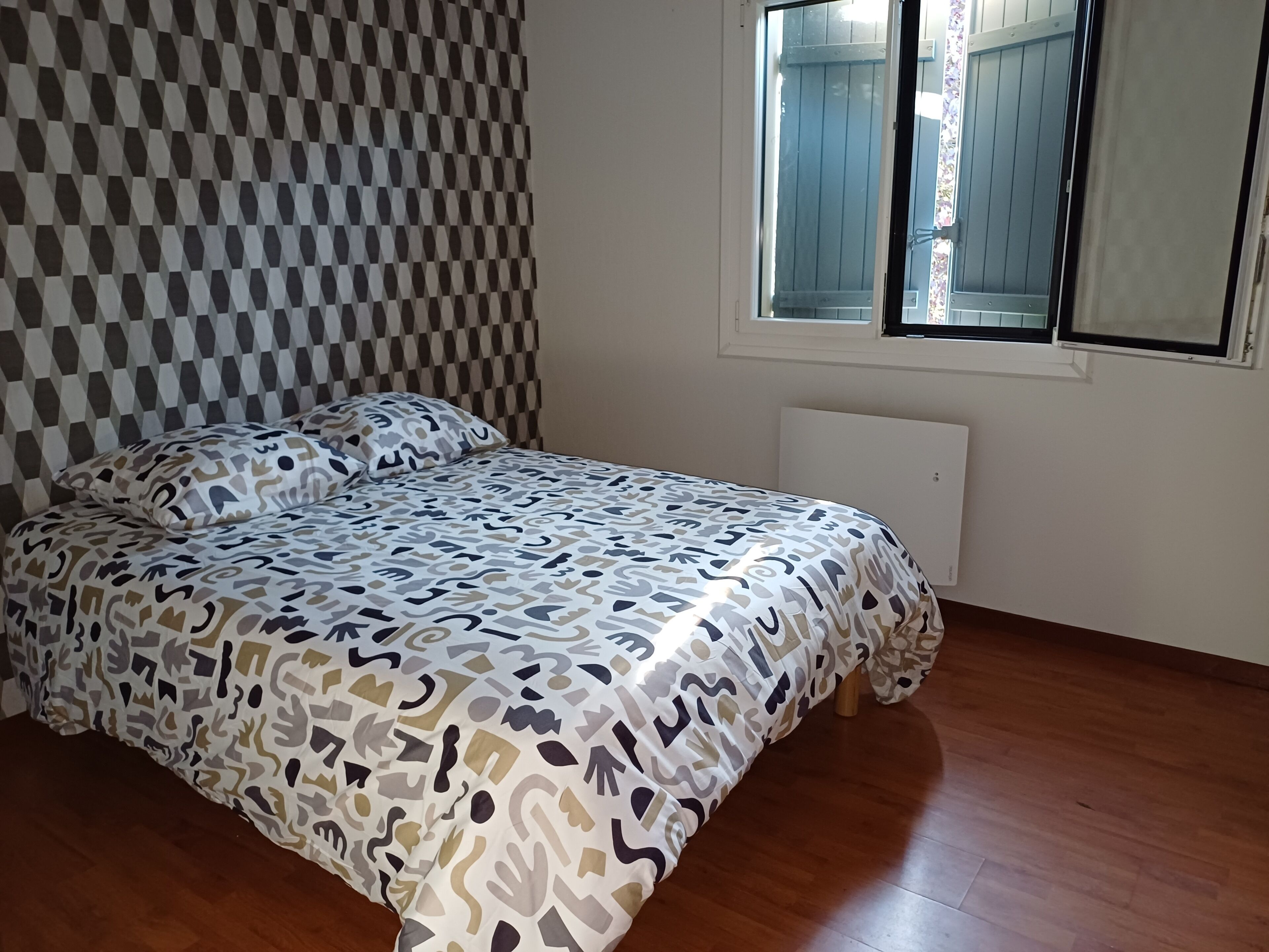 A bedroom with a checkered wall, a patterned bedspread, a window with blinds, and a radiator.