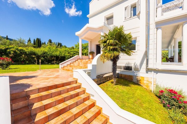 Un bâtiment blanc avec un escalier menant à un porche abrité, entouré d’un jardin bien entretenu et sous un ciel bleu dégagé.