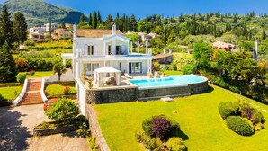 A two-story house with a swimming pool, surrounded by well-maintained gardens and a mountainous backdrop.