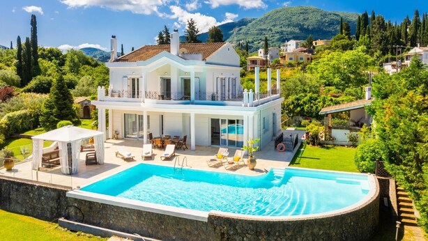 A two-story house with a swimming pool, surrounded by lush greenery and a mountainous backdrop.
