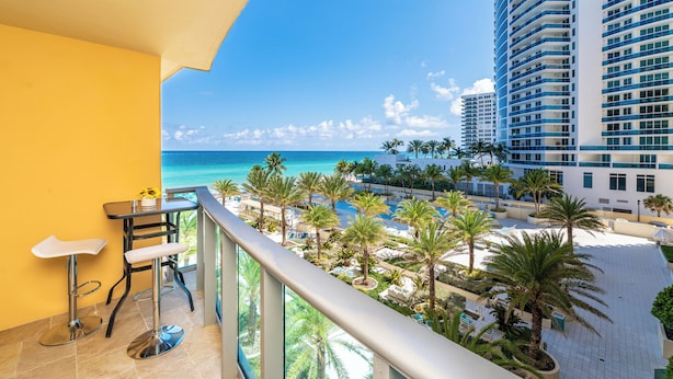 A balcony with a view of the ocean and palm trees, a dining area with two bar stools, and a high-rise building in the background. - 1BD/1.5BTH BeachFront Resort Style. Gym, WiFi, Free Parking, Jacuzzi, GameRoom. (Hollywood)